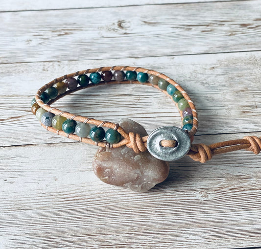 A wrap ankle bracelet featuring multi-color round beads and tan leather. The beads range in color from lavender, burgundy, amber to shades of green. The anklet has an oval silver-toned button with a swirl design as the clasp. The design can wrap around the ankle one time. The bracelet is displayed resting on a light-colored stone on a light wood surface.