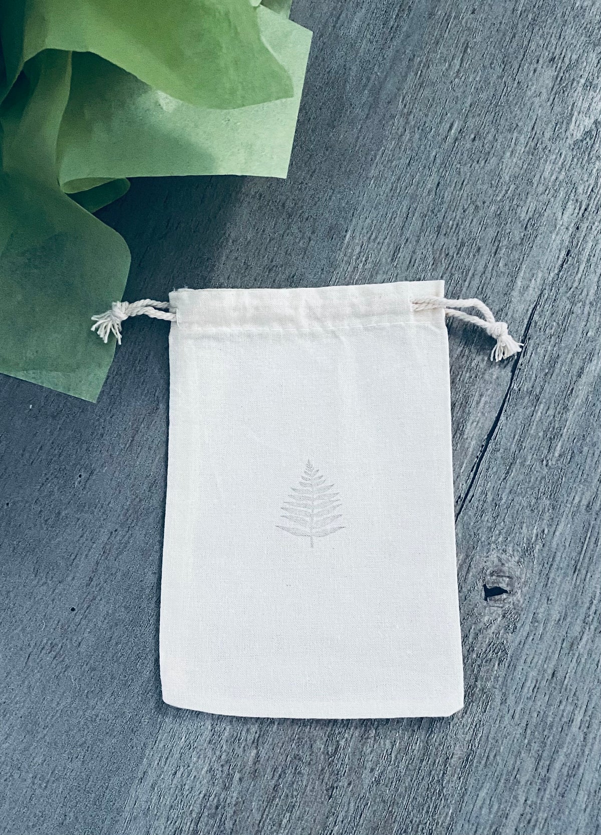 A small off-white fabric drawstring bag with a subtle stamped fern leaf design. The bag is displayed on a gray wood surface. Green tissue paper is visible in the background.