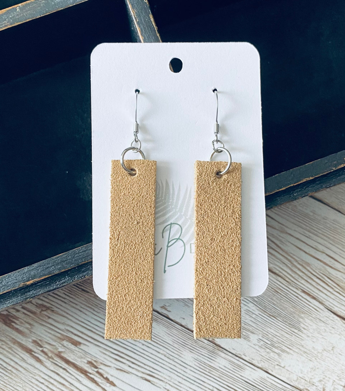 A pair of long rectangular tan suede earrings with silver earring wires. The earrings are displayed on a white branded display card with a black display box and light wooden background.