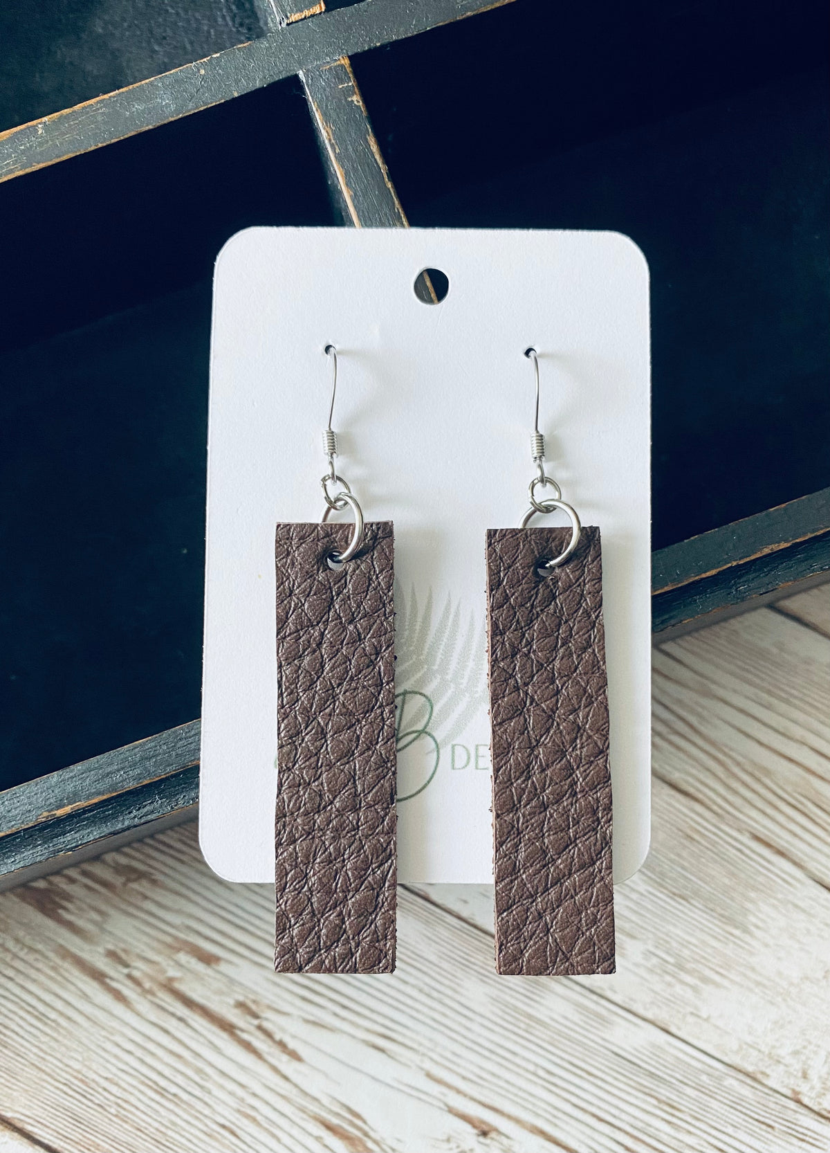 A pair of long rectangular brown leather earrings with silver earring wires. The earrings are displayed on a white branded display card with a black display box and light wooden background.