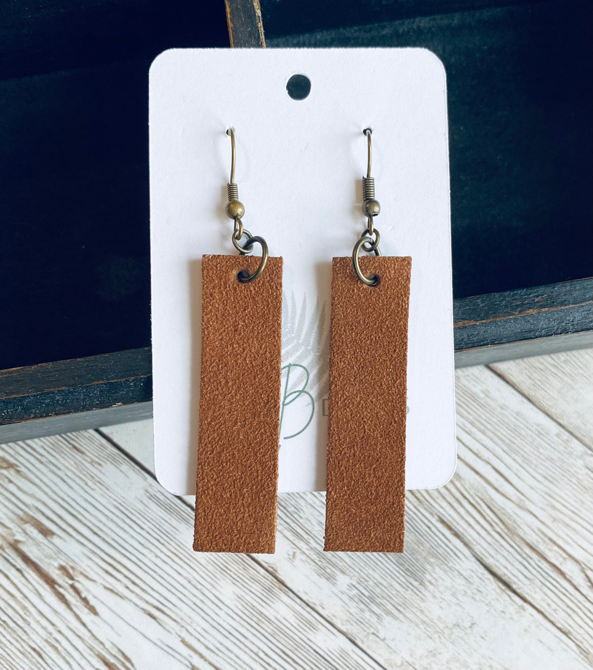 A pair of long rectangular medium brown suede earrings with bronze earring wires. The earrings are displayed on a white branded display card with a black display box and light wooden background.
