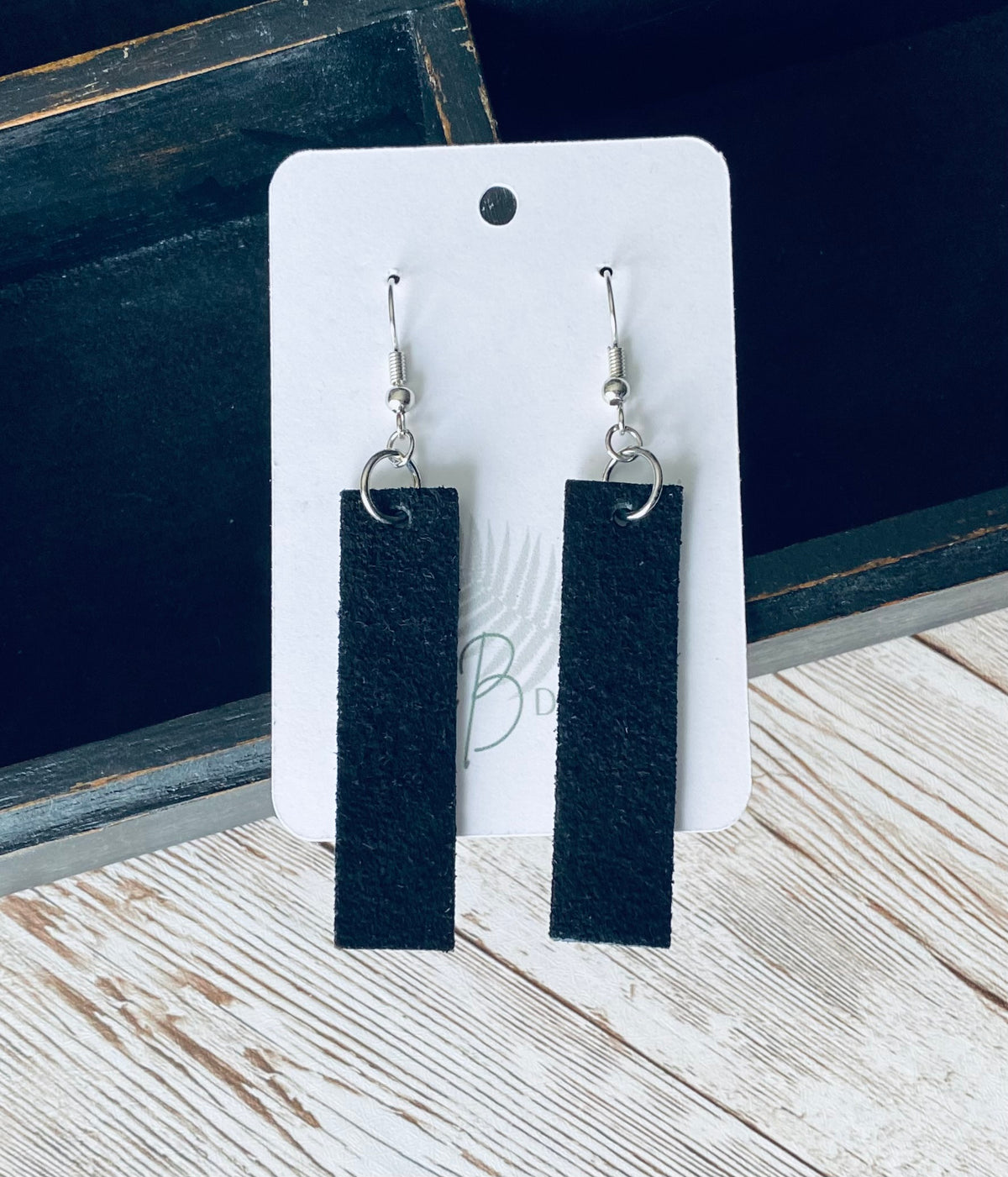 A pair of long rectangular black suede earrings with silver earring wires. The earrings are displayed on a white branded display card with a black display box and light wooden background.