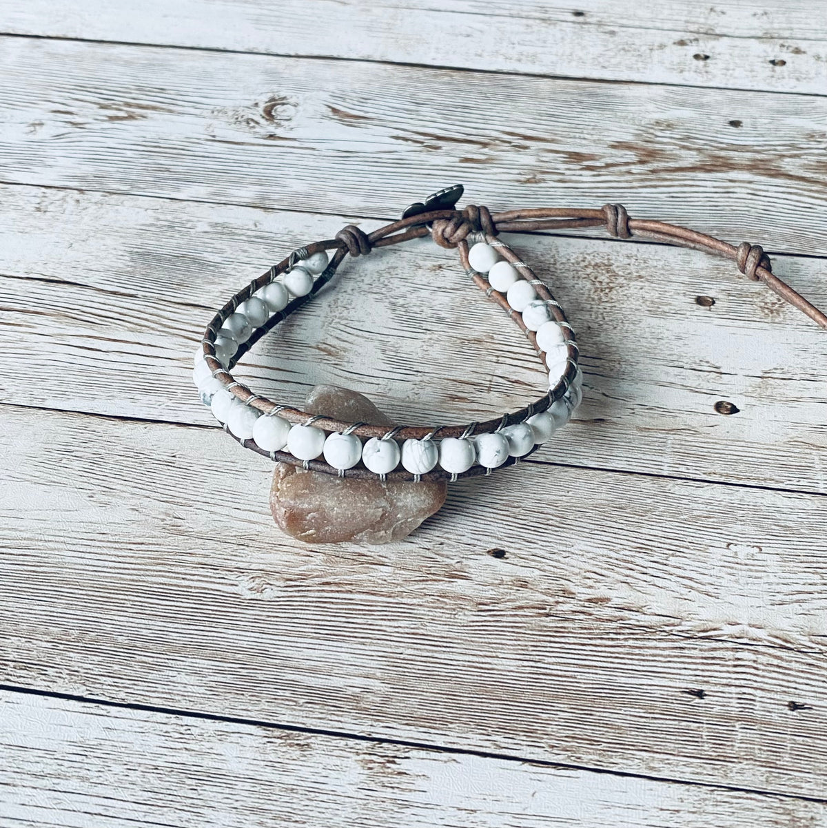 A wrap ankle bracelet featuring white gemstone beads and natural gray leather. The anklet has a gray button clasp.  The design can wrap around the ankle one time.  The bracelet is displayed resting on a light-colored stone on a light wood surface.