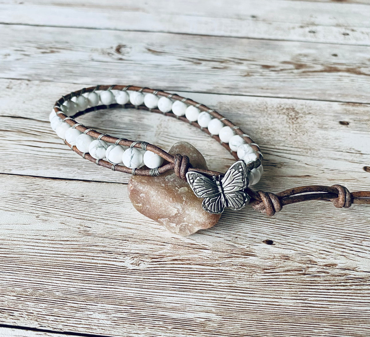 A wrap ankle bracelet featuring white gemstone beads and natural gray leather. The anklet has a gray butterfly button clasp.  The design can wrap around the ankle one time.  The anklet is displayed resting on a light-colored stone on a light wood surface.