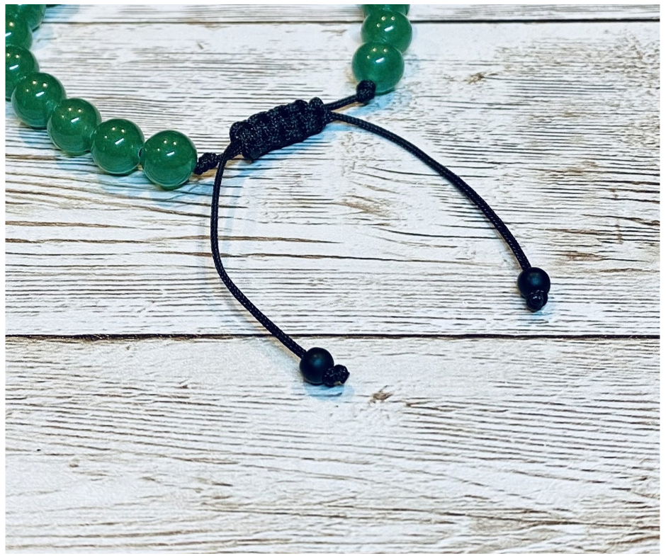 A close-up of a bracelet with round green aventurine gemstone beads, featuring the black adjustable cord with round matte-black bead tips, displayed on a light weathered wood surface.
