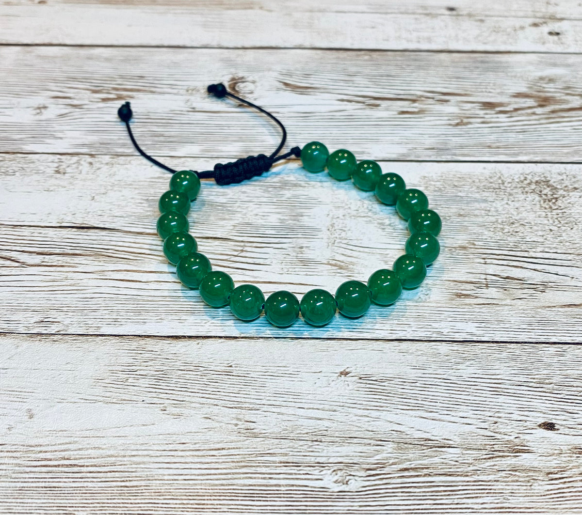 A beaded bracelet made with round green aventurine gemstone beads on an adjustable black nylon cord, with small beads at the ends of the nylon cord.   It is displayed on a light wood background.