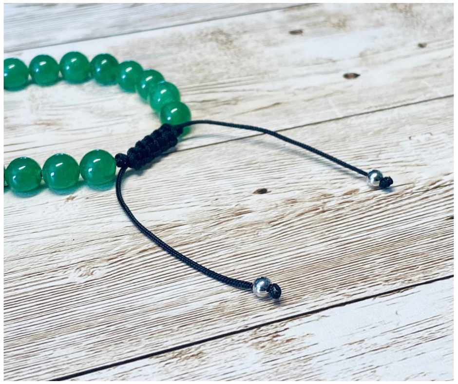 A close-up of a bracelet with round green aventurine gemstone beads, featuring the black adjustable cord with round silver-toned metal bead tips, displayed on a light weathered wood surface.