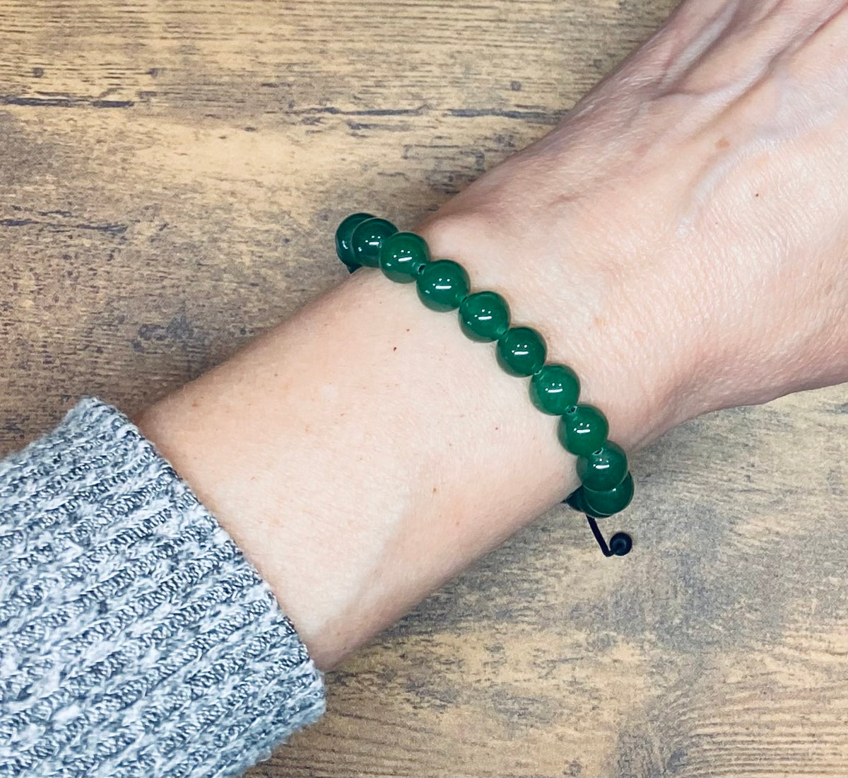 A green aventurine gemstone beaded bracelet worn on a person's wrist, shown against a wooden tabletop background with a gray knit sleeve visible.