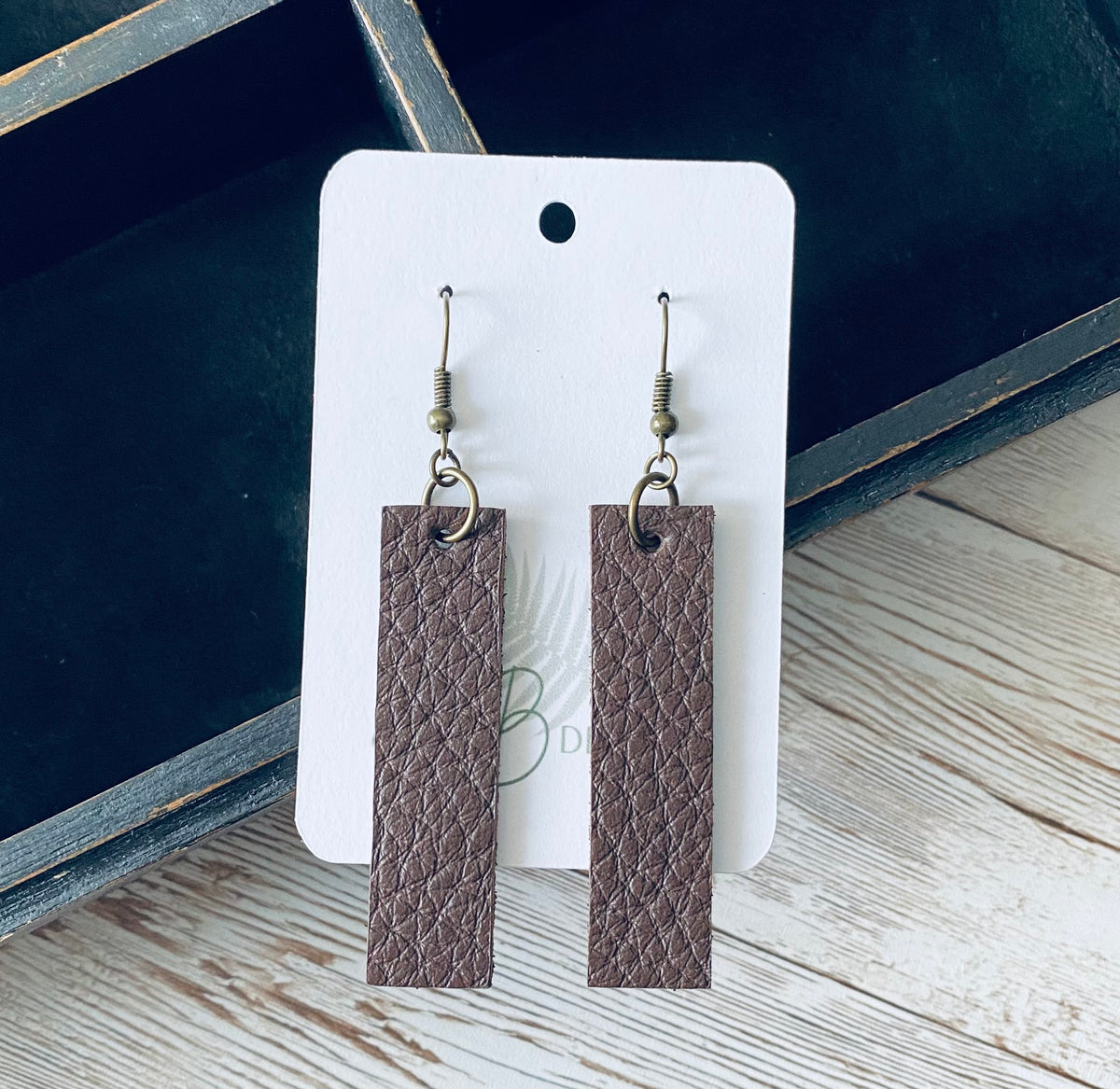 A pair of long rectangular brown leather earrings with bronze earring wires. The earrings are displayed on a white branded display card with a black display box and light wooden background.