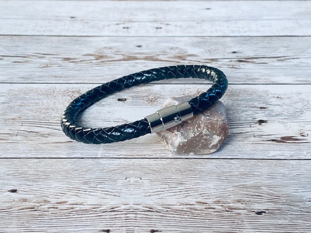 A black braided leather bracelet with a silver magnetic clasp, resting on a small natural stone against a light gray wooden background.