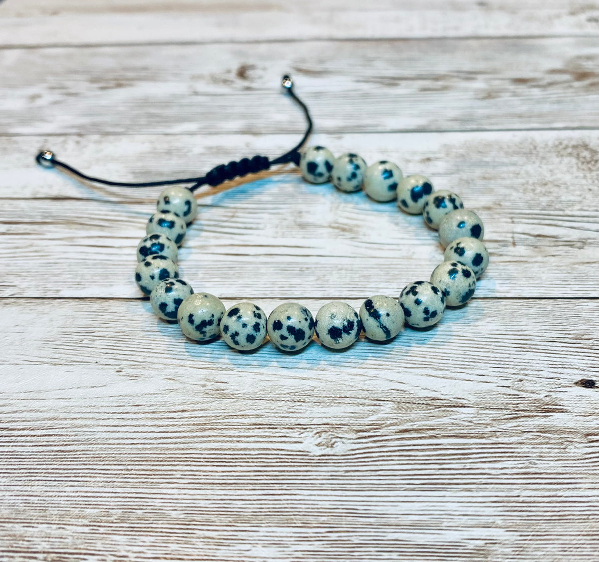 A beaded bracelet made with round beige and black speckled dalmatian jasper gemstone beads on an adjustable black nylon cord, with small beads at the ends of the nylon cord.  It is displayed on a light wood background.