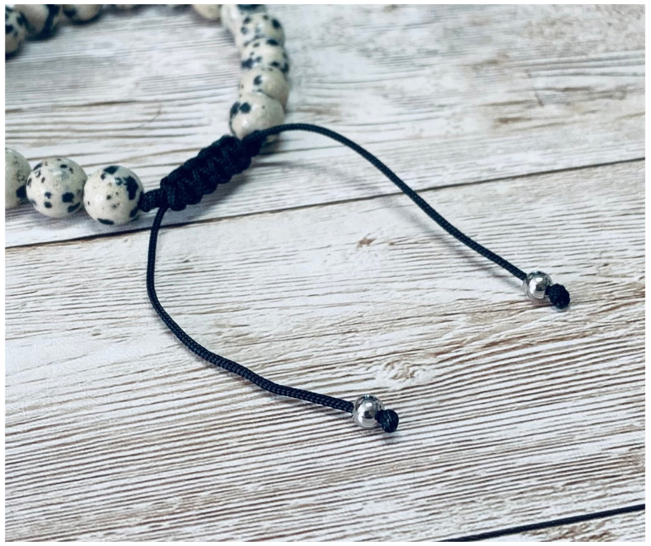 A close-up of a bracelet with round beige with black speckled dalmatian jasper gemstone beads, featuring the black adjustable cord with round silver-toned metal bead tips, displayed on a light weathered wood surface.