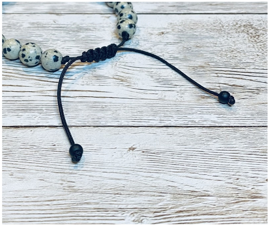 A close-up of a bracelet with round beige with black speckled dalmatian jasper gemstone beads, featuring the black adjustable cord with round matte-black bead tips, displayed on a light weathered wood surface.
