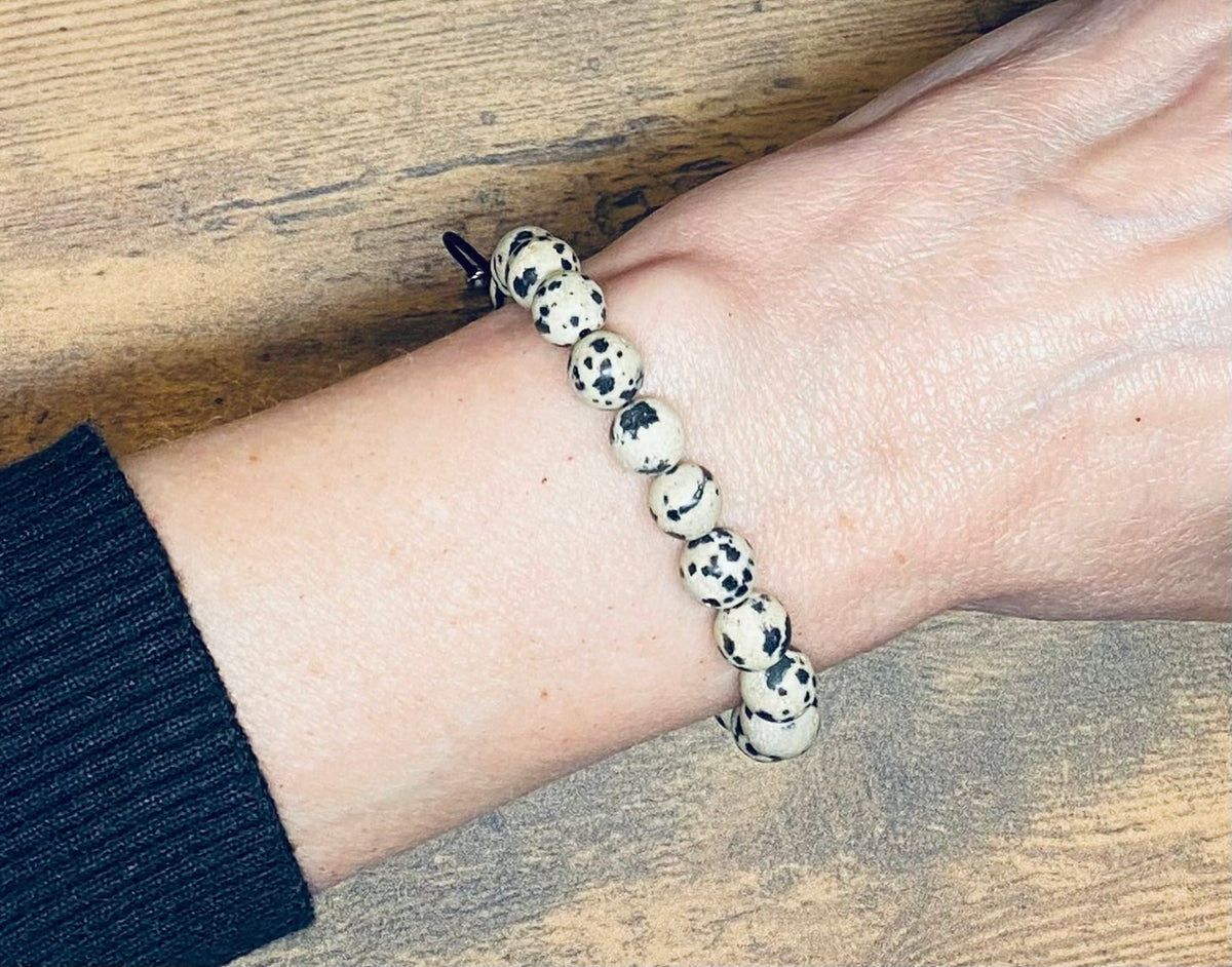 A beige and black speckled dalmatian jasper gemstone beaded bracelet worn on a person's wrist, shown against a wooden tabletop background with a black knit sleeve visible.