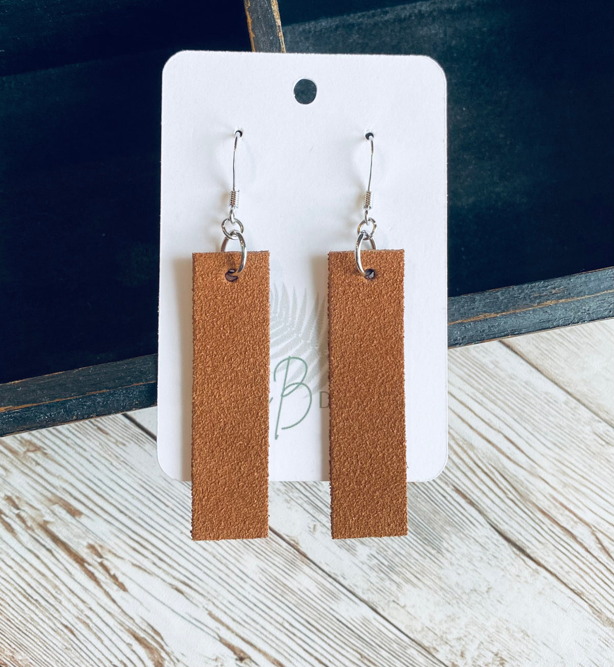 A pair of long rectangular medium brown suede earrings with silver earring wires. The earrings are displayed on a white branded display card with a black display box and light wooden background.