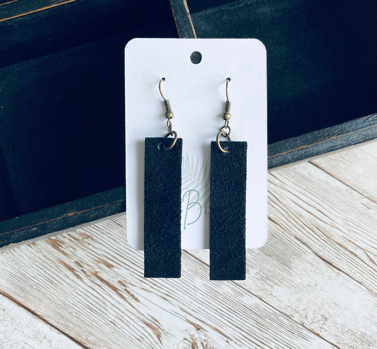 A pair of long rectangular black suede earrings with bronze earring wires. The earrings are displayed on a white branded display card with a black display box and light wooden background.
