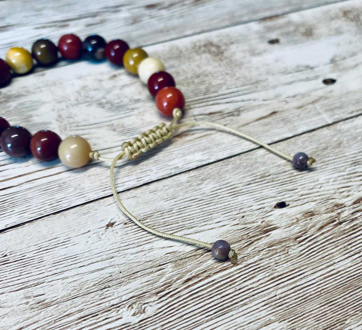 A close-up of a bracelet with mookaite jasper beads in warm tones of burgundy, mustard yellow, cream and shades of purple.  It is featuring the cream tone adjustable cord with lavender tone fancy jasper bead tips, displayed on a light weathered wood surface.