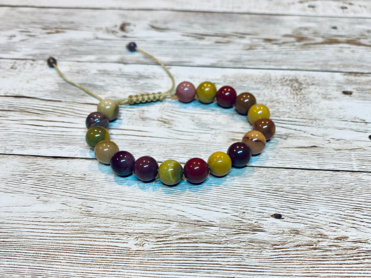 A beaded bracelet made with round Mookaite jasper gemstone beads on an adjustable cream tone nylon cord, with small beads at the ends of the nylon cord.    The beads include warm tones of burgundy, mustard yellow, cream and shades of purple.  It is displayed on a light wood background.