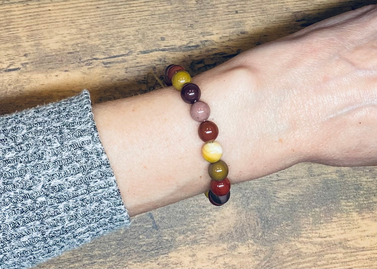 A bracelet with mookaite jasper gemstone beads in warm tones of burgundy, mustard yellow, cream and shades of purple.  It is displayed on a person's wrist, shown against a wooden tabletop background with a gray knit sleeve visible.