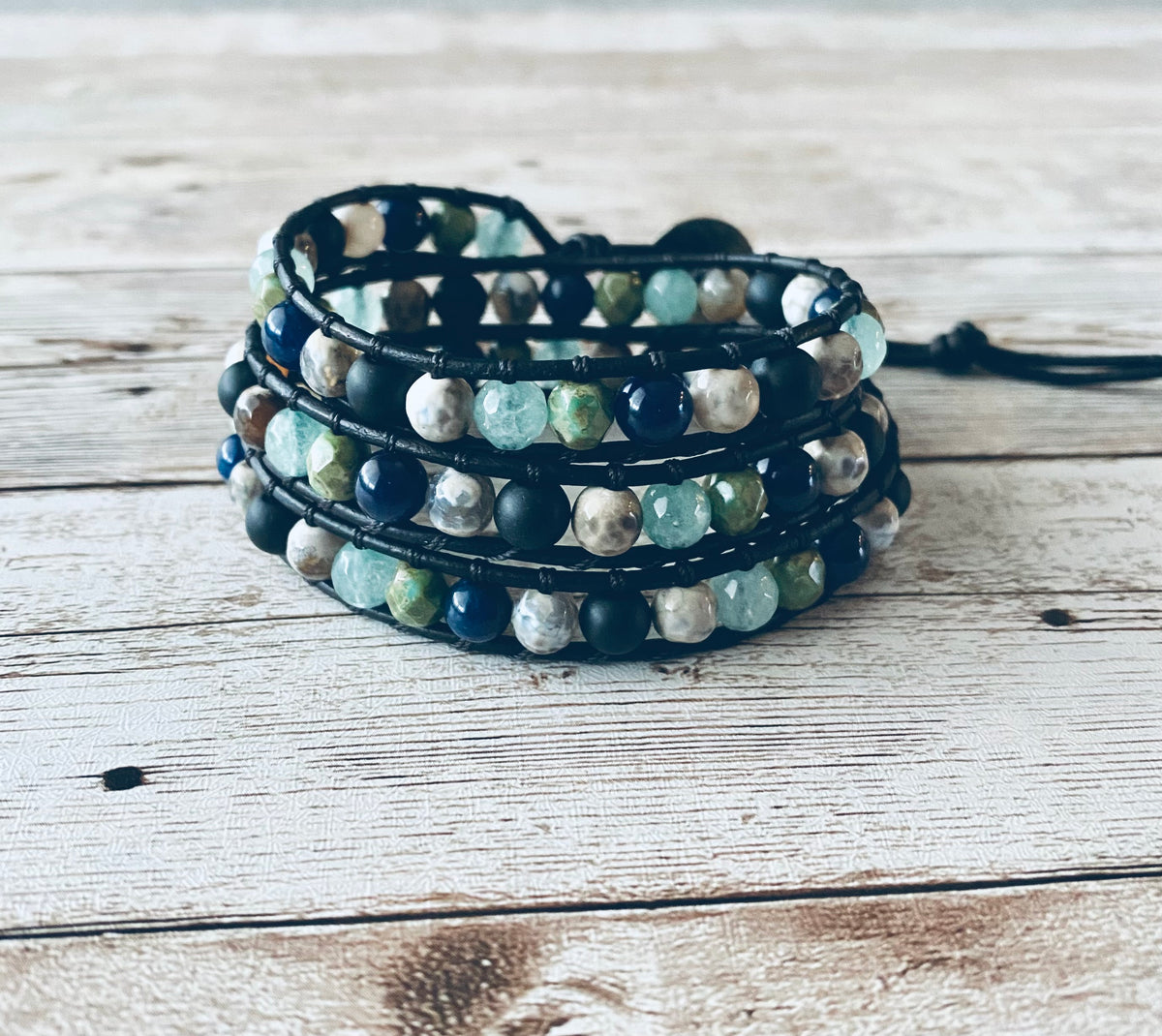 A wrap bracelet featuring navy, aqua blue, green and black beads. The beads are strung on black leather cord, creating a layered effect. The bracelet is designed to wrap around the wrist three times.  It is displayed on a light wood background. 