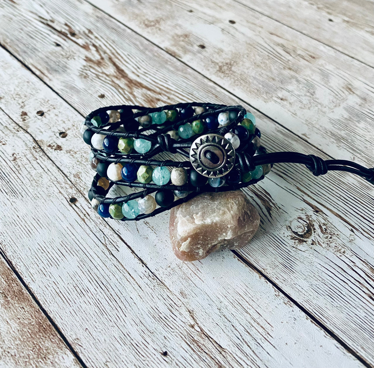 A wrap bracelet featuring navy, aqua blue, green and black beads. The beads are strung on black leather cord and have a silver button as the closure.  The bracelet is designed to wrap around the wrist three times.  It is displayed resting on a light-colored stone on a light wood background. 