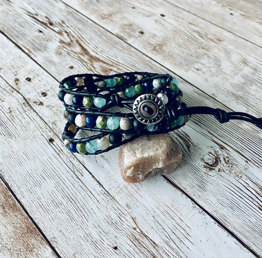 A wrap bracelet featuring navy, aqua blue, green and black beads. The beads are strung on black leather cord and have a silver button as the closure.  The bracelet is designed to wrap around the wrist three times.  It is displayed resting on a light-colored stone on a light wood background. 