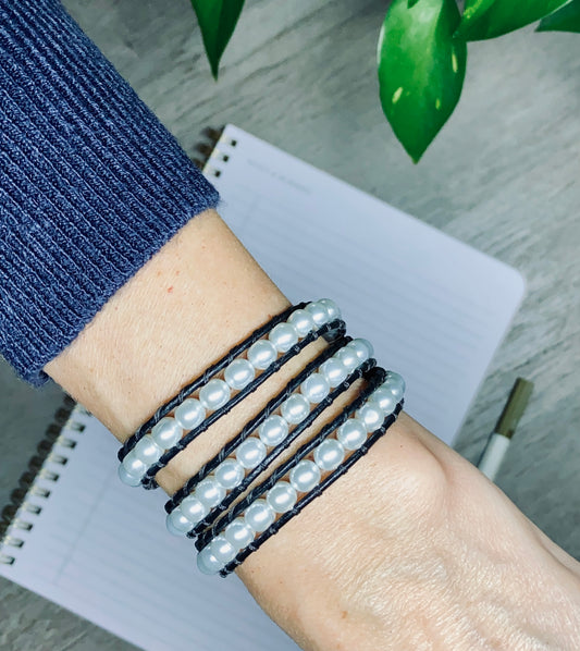 Close-up of a wrist wearing triple wrap pearl and black leather bracelet with a notebook and plant in the background.