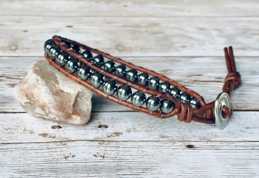 A wrap bracelet featuring round dark gray gemstone beads.  The beads are strung on a natural antique brown leather cord with a decorative antique silver tone button clasp and is designed to wrap one time around the wrist.  The bracelet is displayed resting on a light-colored stone on a light wood surface.