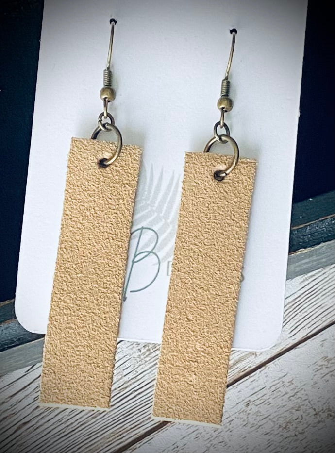 A pair of long rectangular light tan suede earrings with bronze earring wires.  The earrings are displayed on a white branded display card with a black display box and light wooden background.