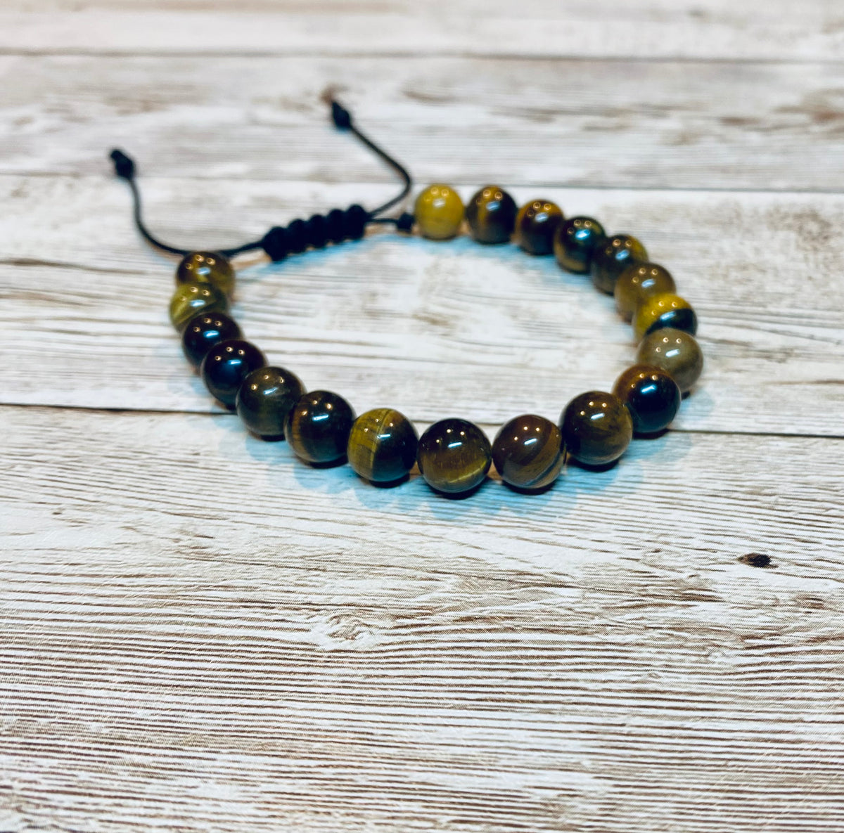 A beaded bracelet made with round brown and gold tiger's eye gemstone beads on an adjustable black nylon cord, with small beads at the ends of the nylon cord.  It is displayed on a light wood background.