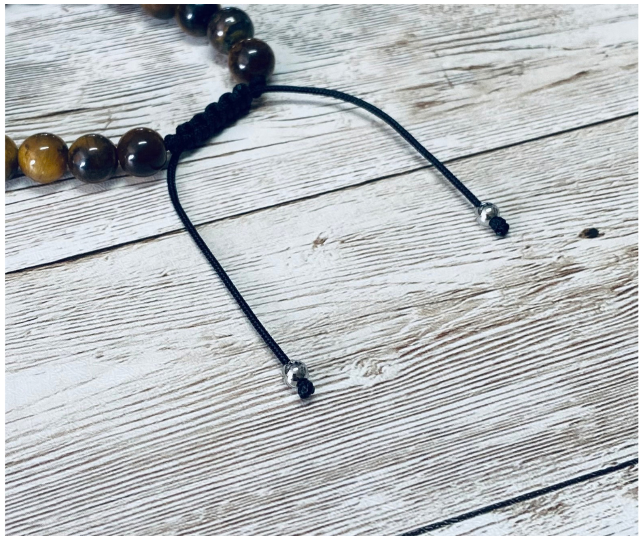 A close-up of a bracelet with brown and gold tiger's eye gemstone beads, featuring the black adjustable cord with round silver-toned metal bead tips, displayed on a light weathered wood surface.