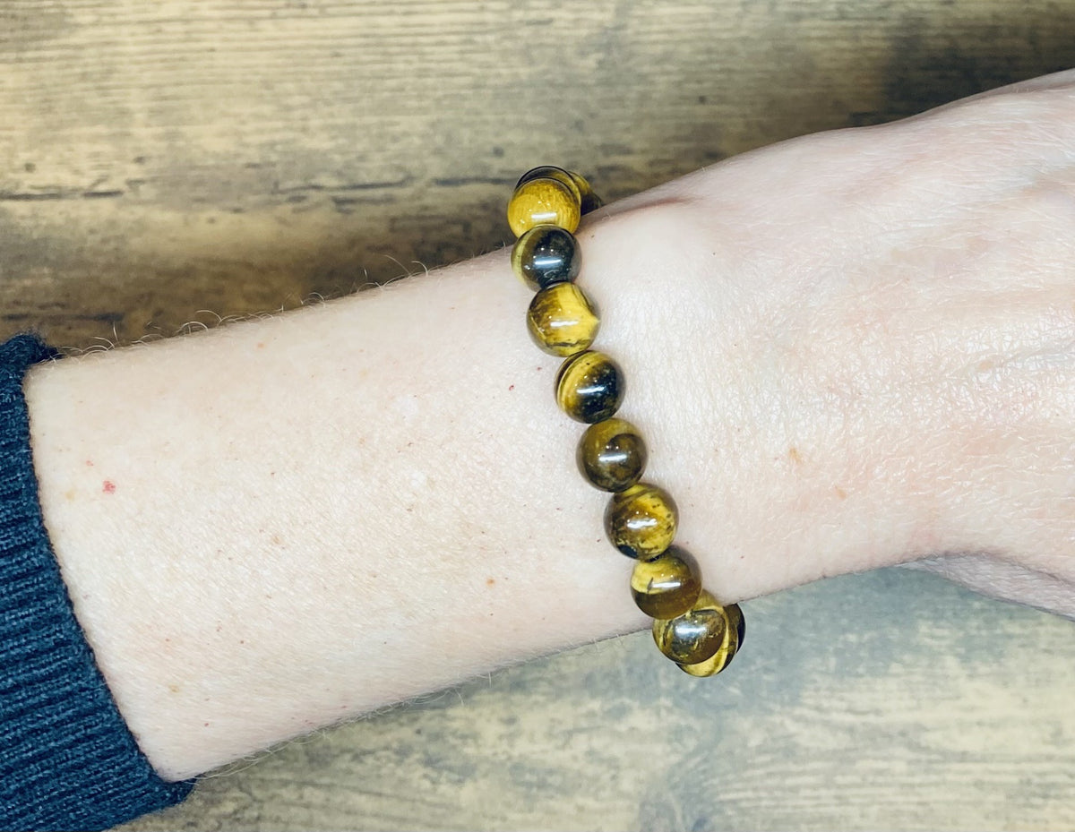 A brown and gold tiger's eye gemstone beaded bracelet worn on a person's wrist, shown against a wooden tabletop background with a black knit sleeve visible.