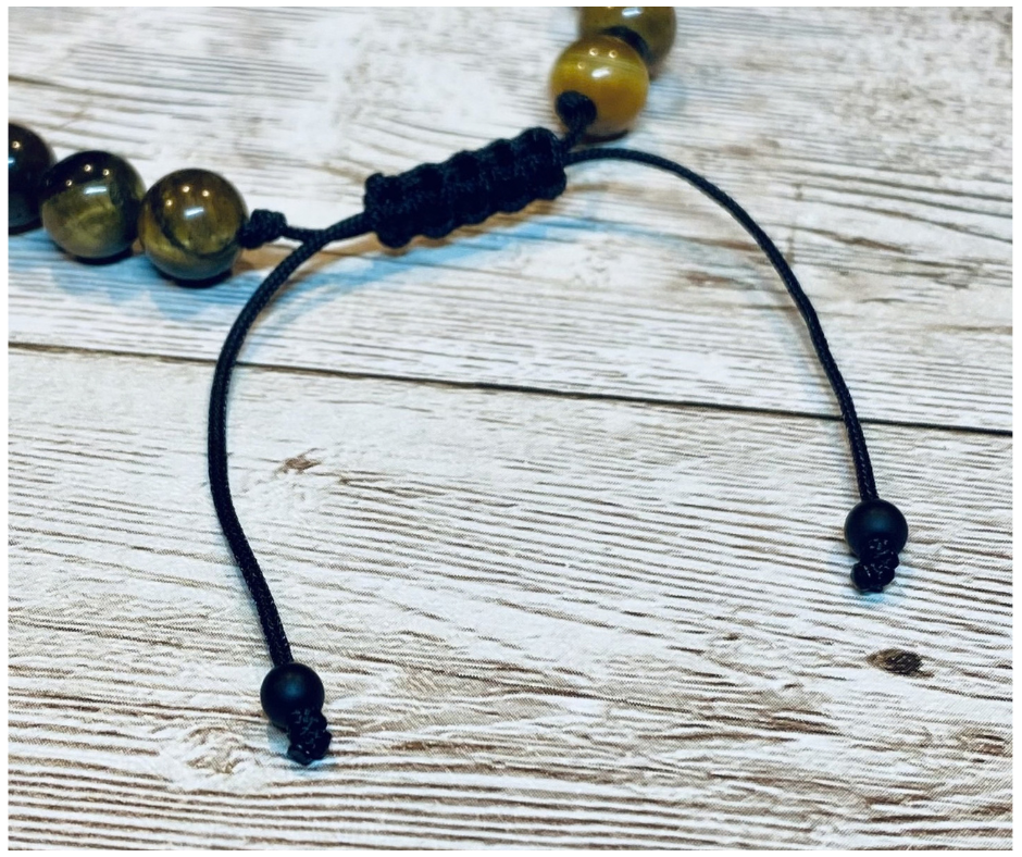 A close-up of a bracelet with round brown and gold tiger's eye gemstone beads, featuring the black adjustable cord with round matte-black bead tips, displayed on a light weathered wood surface.