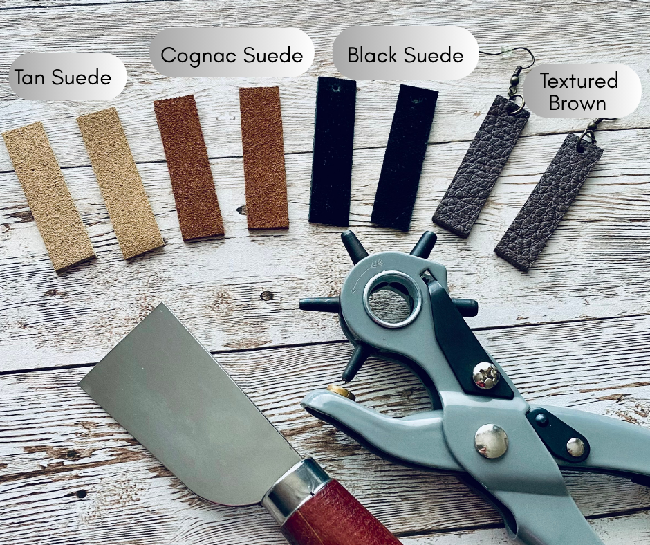 Leather punching and cutting tools with earring materials displayed on a light wooden surface.  The earring materials are labeled with the color and texture of the material, including tan suede, cognac suede, black suede and textured brown.