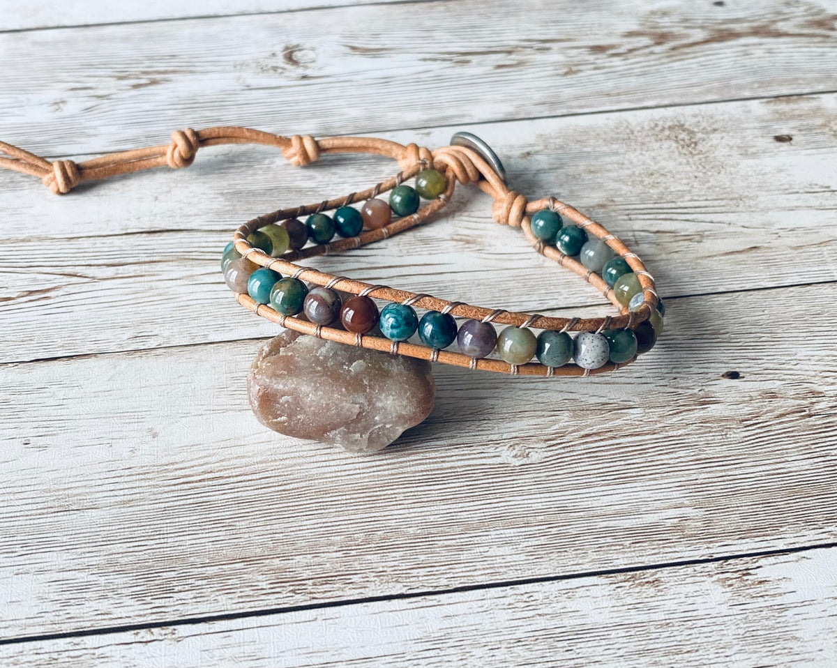 A wrap ankle bracelet featuring multi-color round beads and tan leather. The beads range in color from lavender, burgundy, amber to shades of green.  The anklet has a silver-toned button clasp.  The design can wrap around the ankle one time.  The bracelet is displayed resting on a light-colored stone on a light wood surface.