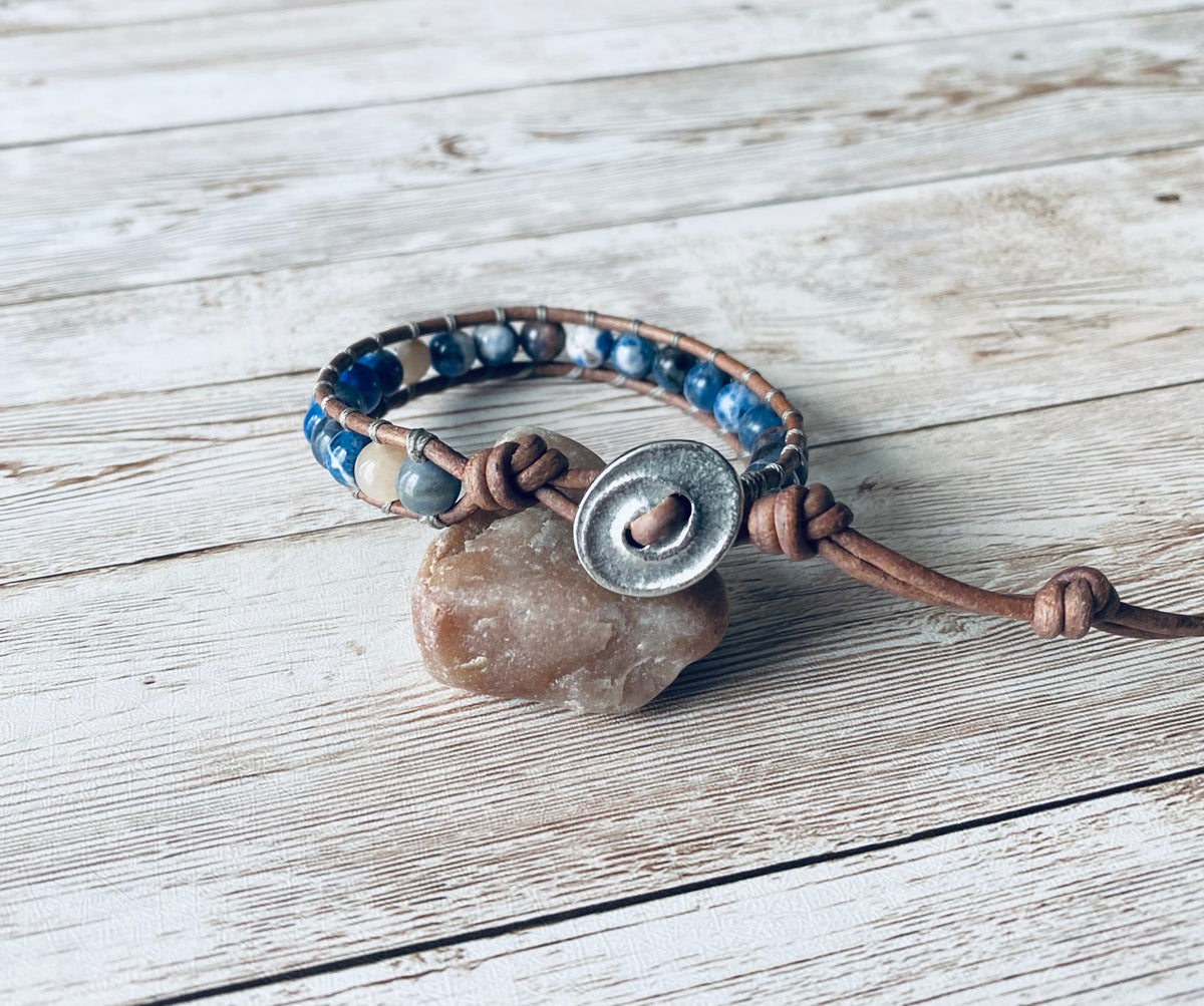 A wrap bracelet featuring blue and white gemstone beads strung on a natural gray leather cord. The bracelet has an oval silver-toned button with a swirl design as the clasp.  It will wrap around the wrist one time.  The bracelet is displayed resting on a light-colored stone on a light wood surface.