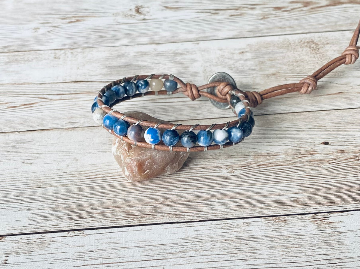 A wrap bracelet featuring blue and white gemstone beads strung on a natural gray leather cord. The bracelet has a silver-tone button clasp and wraps around the wrist one time.  The bracelet is displayed resting on a light-colored stone on a light wood surface.