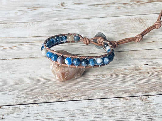 A wrap bracelet featuring blue and white gemstone beads strung on a natural gray leather cord. The bracelet has a silver-tone button clasp and wraps around the wrist one time.  The bracelet is displayed resting on a light-colored stone on a light wood surface.
